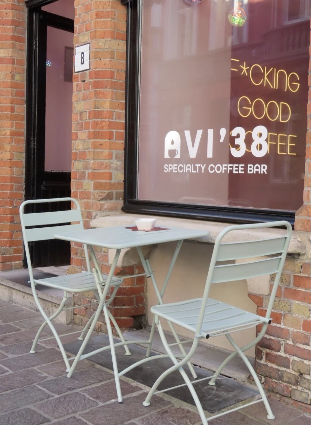 The AVI '38 Specialty Coffee Bar window sign in gold and white lettering, with a small bistro table and two chairs set outside on the brick sidewalk