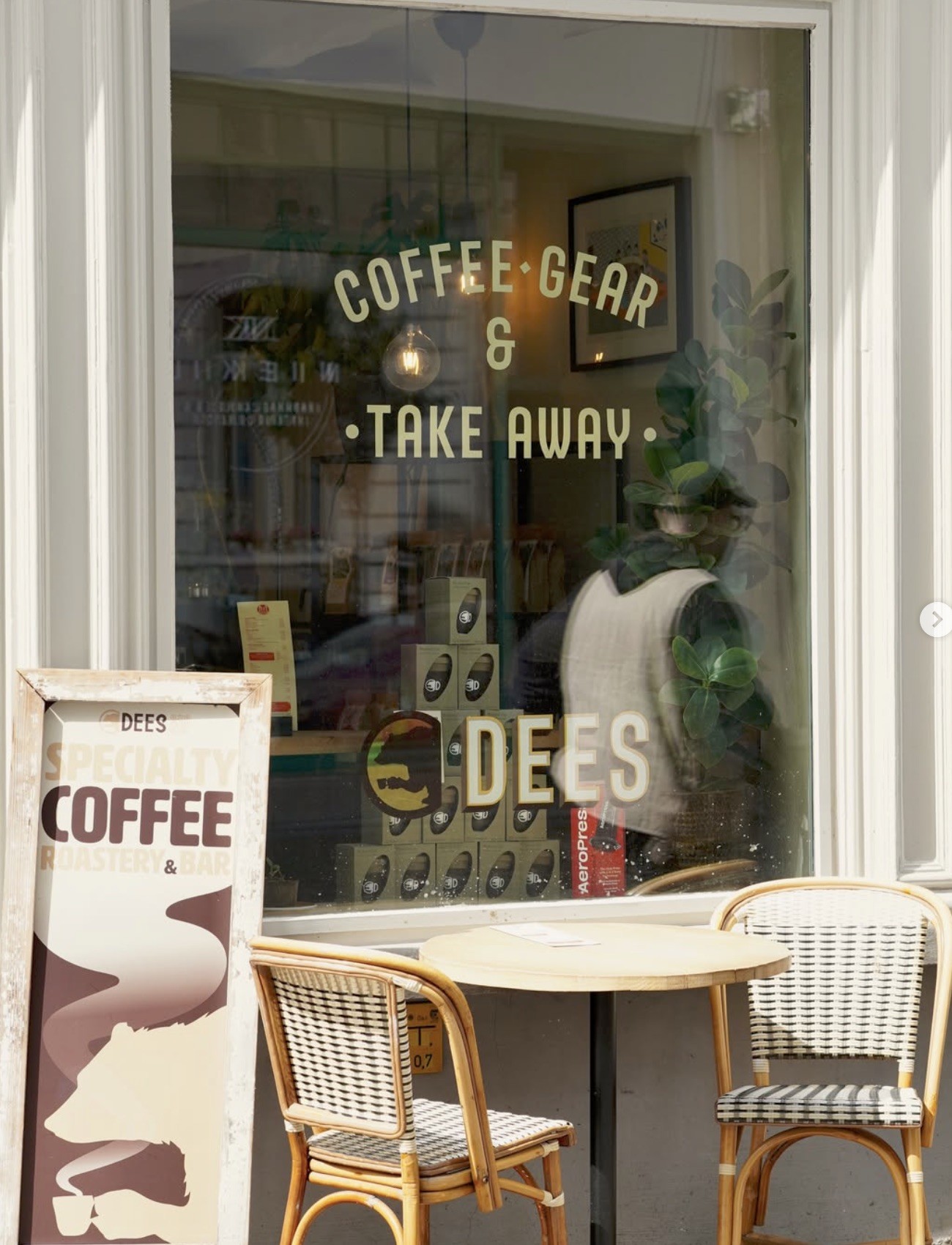 The DEES storefront window with Coffee Gear and Take Away lettering, AeroPress boxes stacked on display, and a bistro table with rattan chairs outside