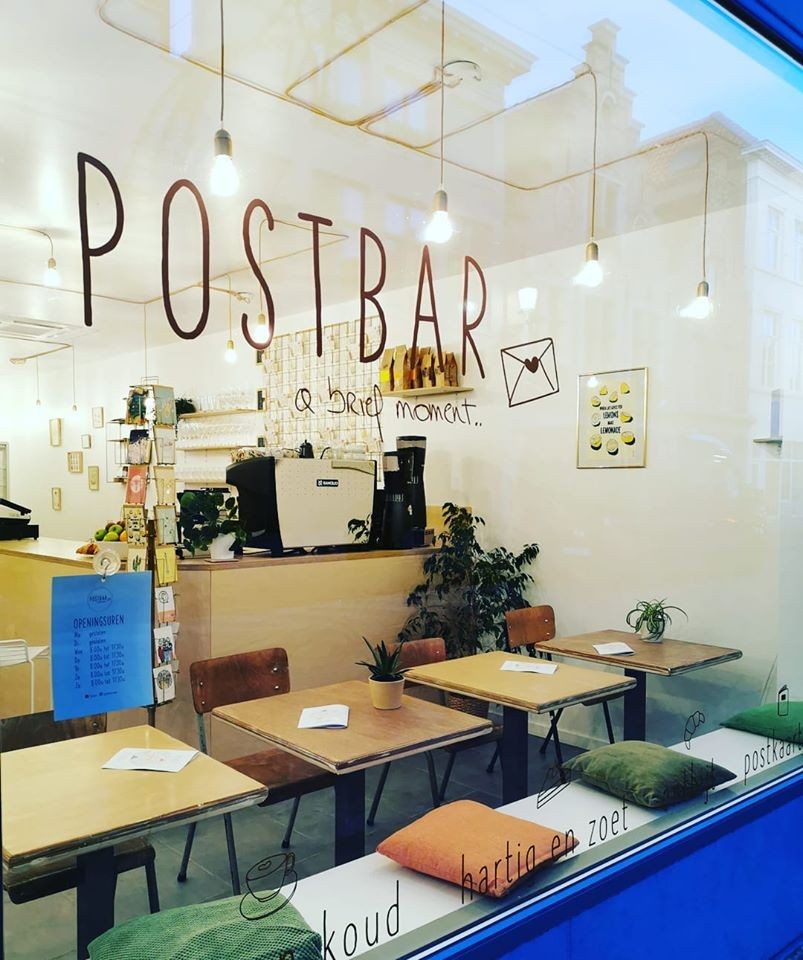Looking through the Postbar window at the hand lettered POSTBAR a brief moment wall logo, an espresso machine on the counter, hanging bulb lights, and small wooden cafe tables with an envelope card rack in the corner