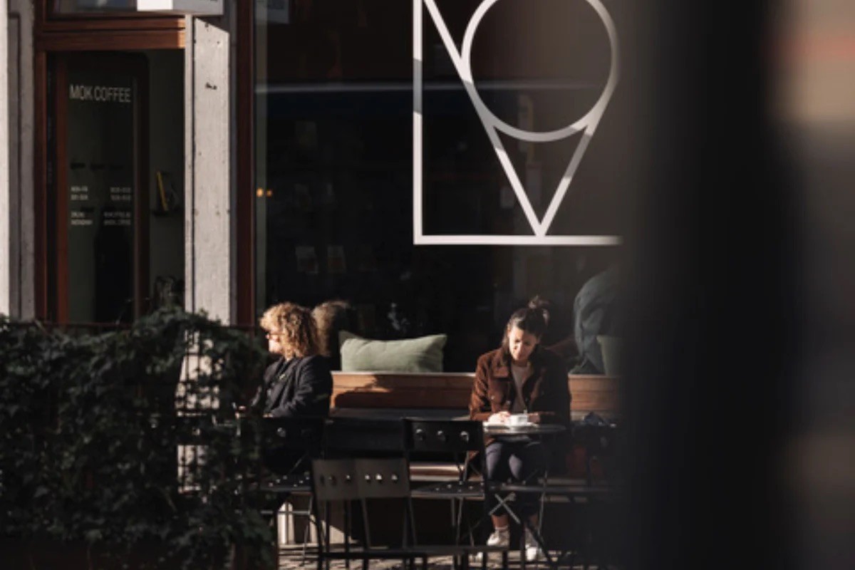 Customers seated at outdoor tables in front of MOK Coffee's storefront window, its geometric logo etched on the glass