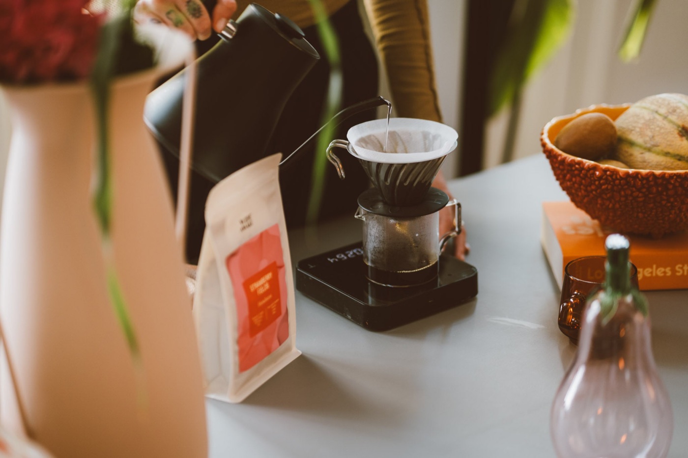 A pour-over brewing on a scale beside a bag of Wide Awake coffee, with fruit and books on the table