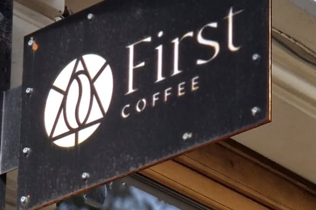 First Coffee's illuminated hanging sign with its circular logo mounted above the shop entrance