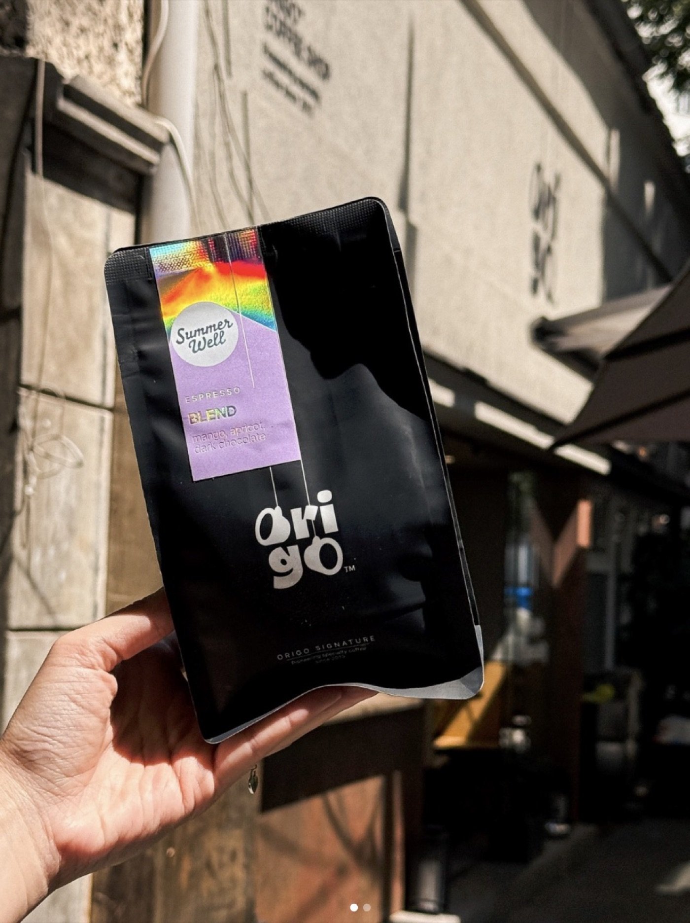 A hand holding a black Origo Coffee retail bag outside the shop