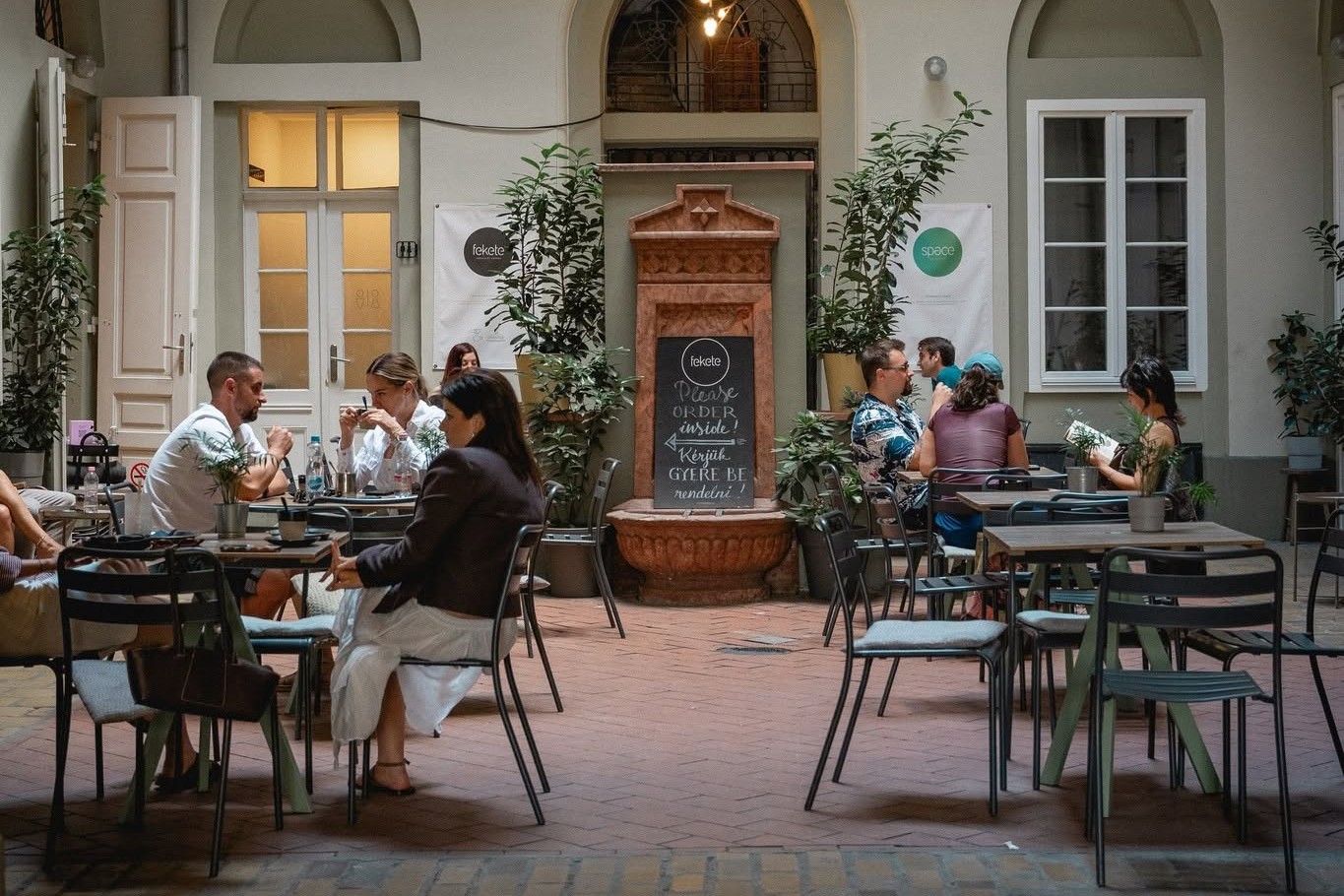 Fekete's courtyard seating area with its terracotta fountain and chalkboard sign, guests seated at tables under the building's arched galleries