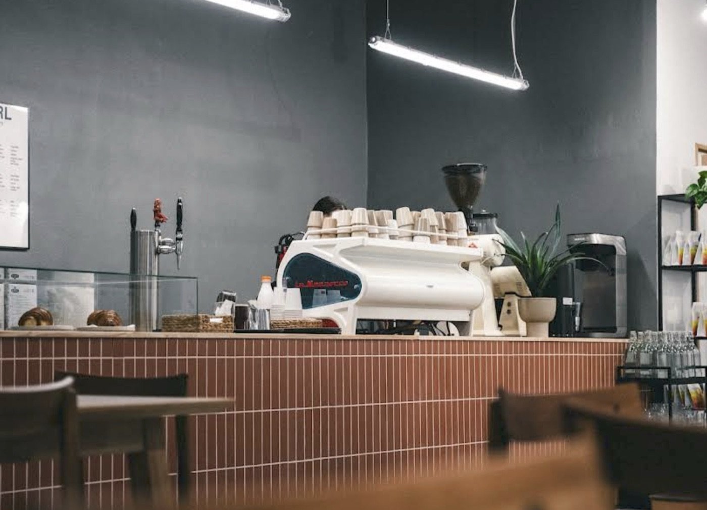 The tiled counter and white La Marzocco espresso machine at Kontakt, cups stacked on top and a drinks fridge behind