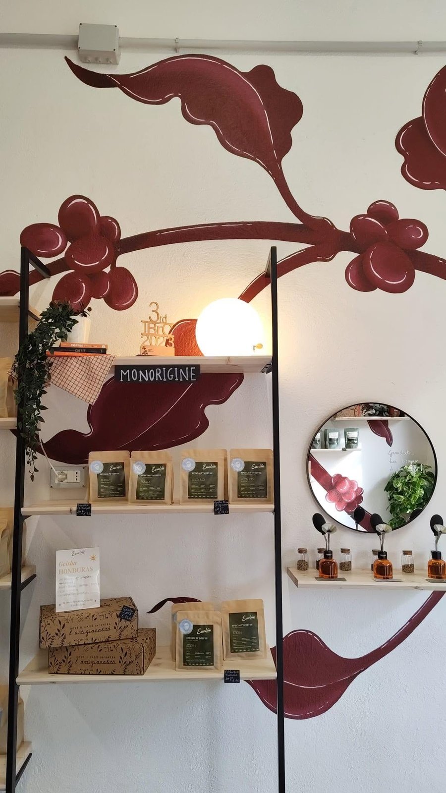 A shelf of bagged Essentzia specialty coffee for retail, labeled by origin, against a wall mural of coffee branches