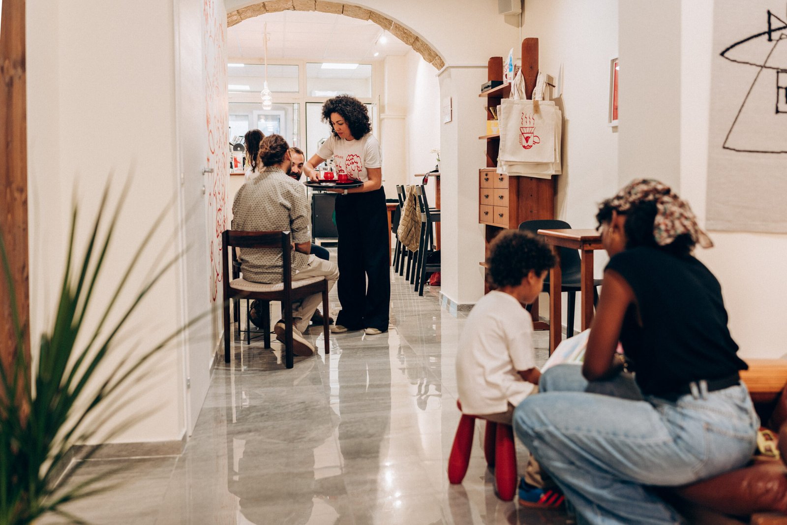 A server carries a tray with a red espresso cup and a glass of soda through L'Atypica's hallway, past seated customers