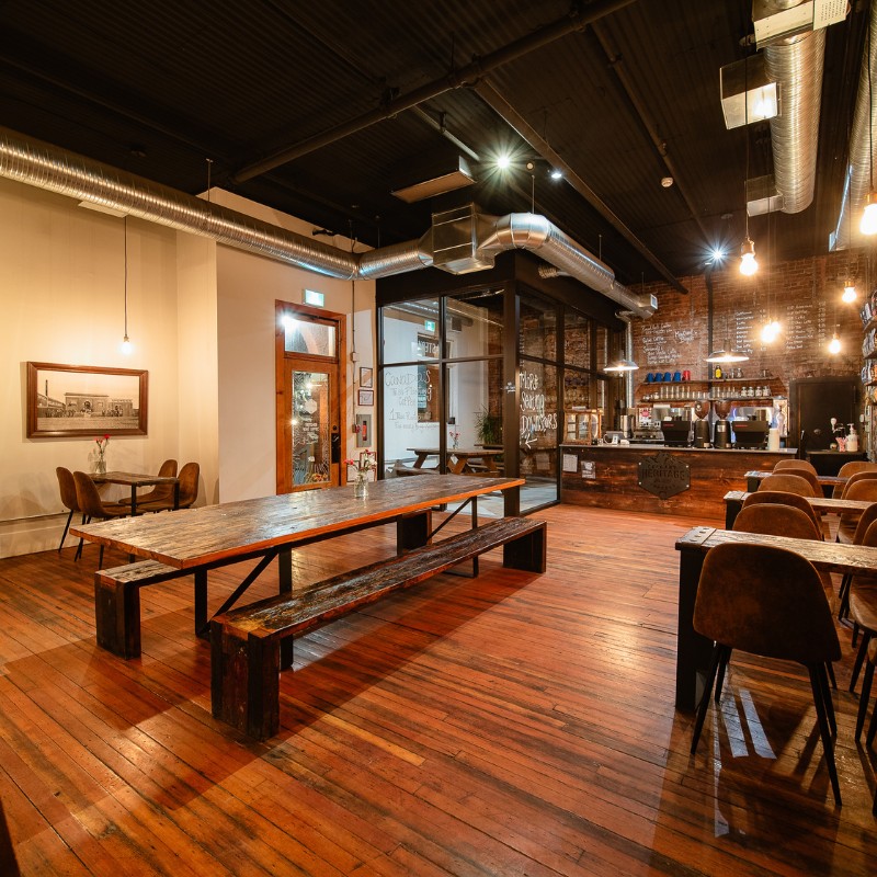 The brick walled cafe floor with communal wood tables, a bench seat, and the espresso bar visible against exposed brick in the C.C. Snowdon building