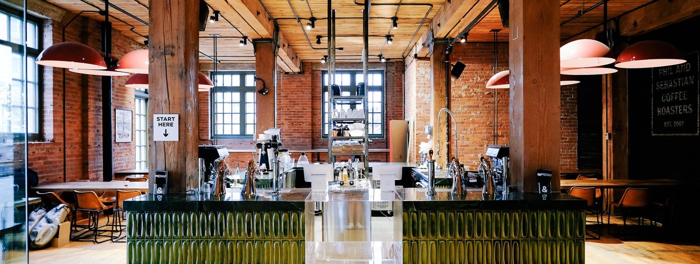 The wide brick and timber cafe floor at the Simmons Building with the espresso bar under red pendant lights and a Phil and Sebastian Coffee Roasters sign visible in the back corner