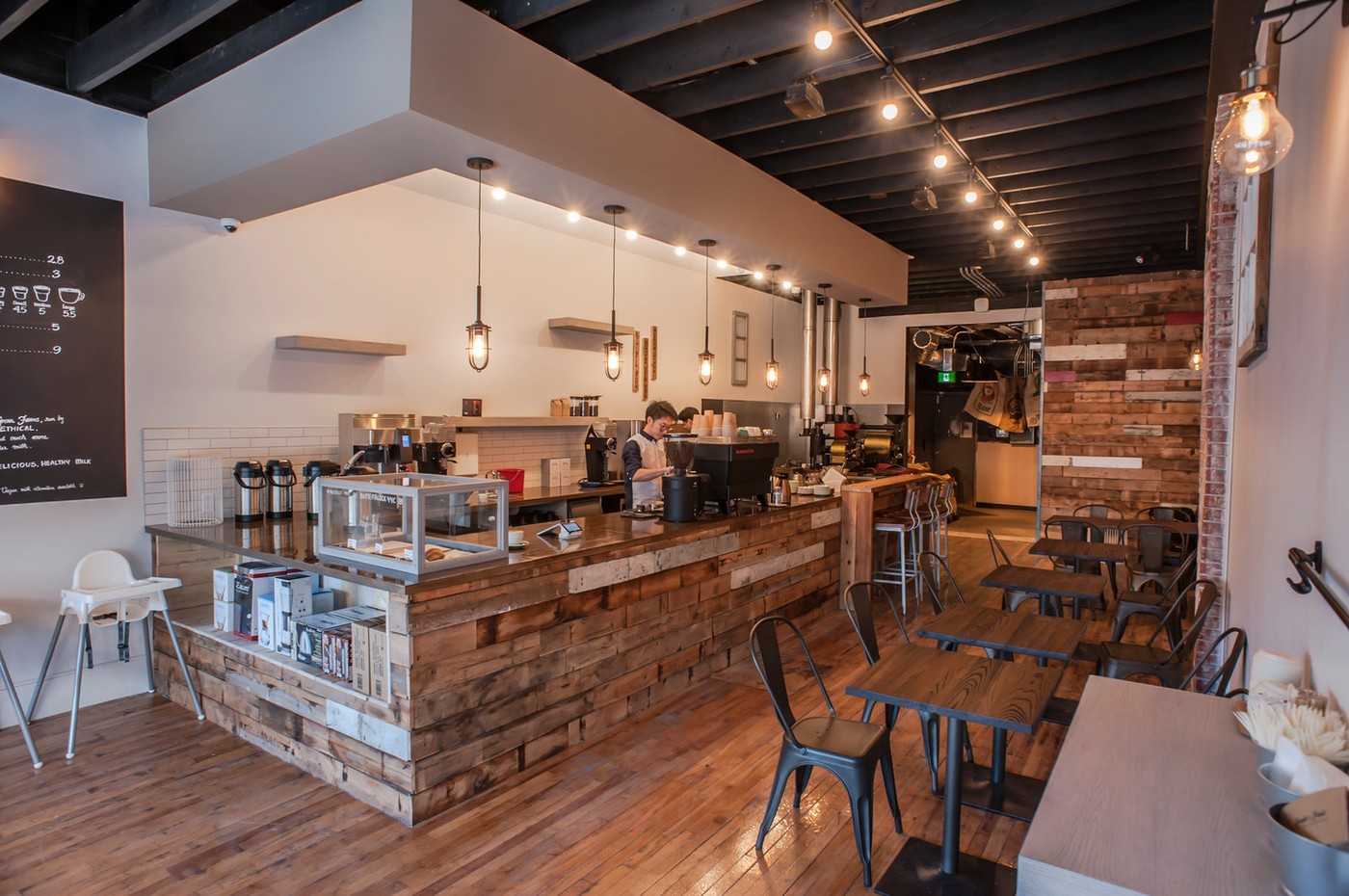 The wood clad counter and espresso bar with a barista grinding coffee under a row of pendant lights, tables and chairs set up along the far wall