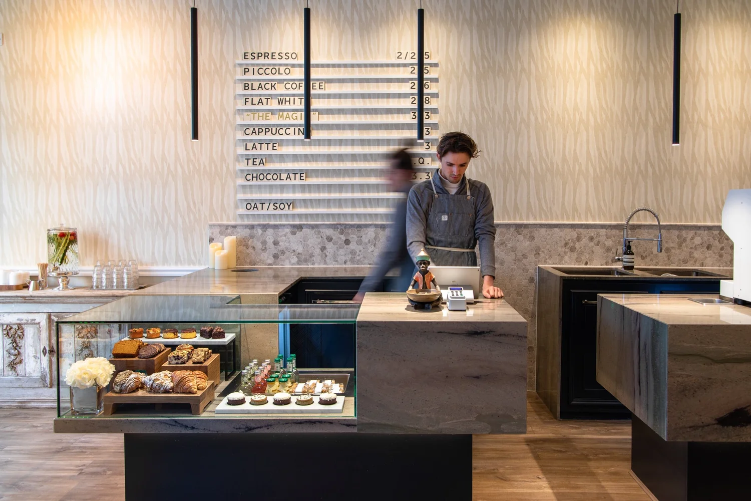 The marble counter at Bould Brothers Coffee, with a hand lettered menu board listing espresso, piccolo, flat white and cappuccino, a glass pastry case of croissants and cakes, and a barista at work