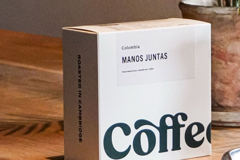 A Coffee World retail bag reading Roasted in Cambridge, Colombia Manos Juntas, standing on a wooden table beside a potted palm