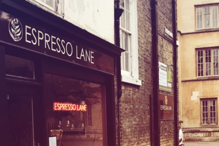 The narrow brick shopfront of Espresso Lane on Botolph Lane, with ESPRESSO LANE lettering above the window and a matching neon sign glowing inside