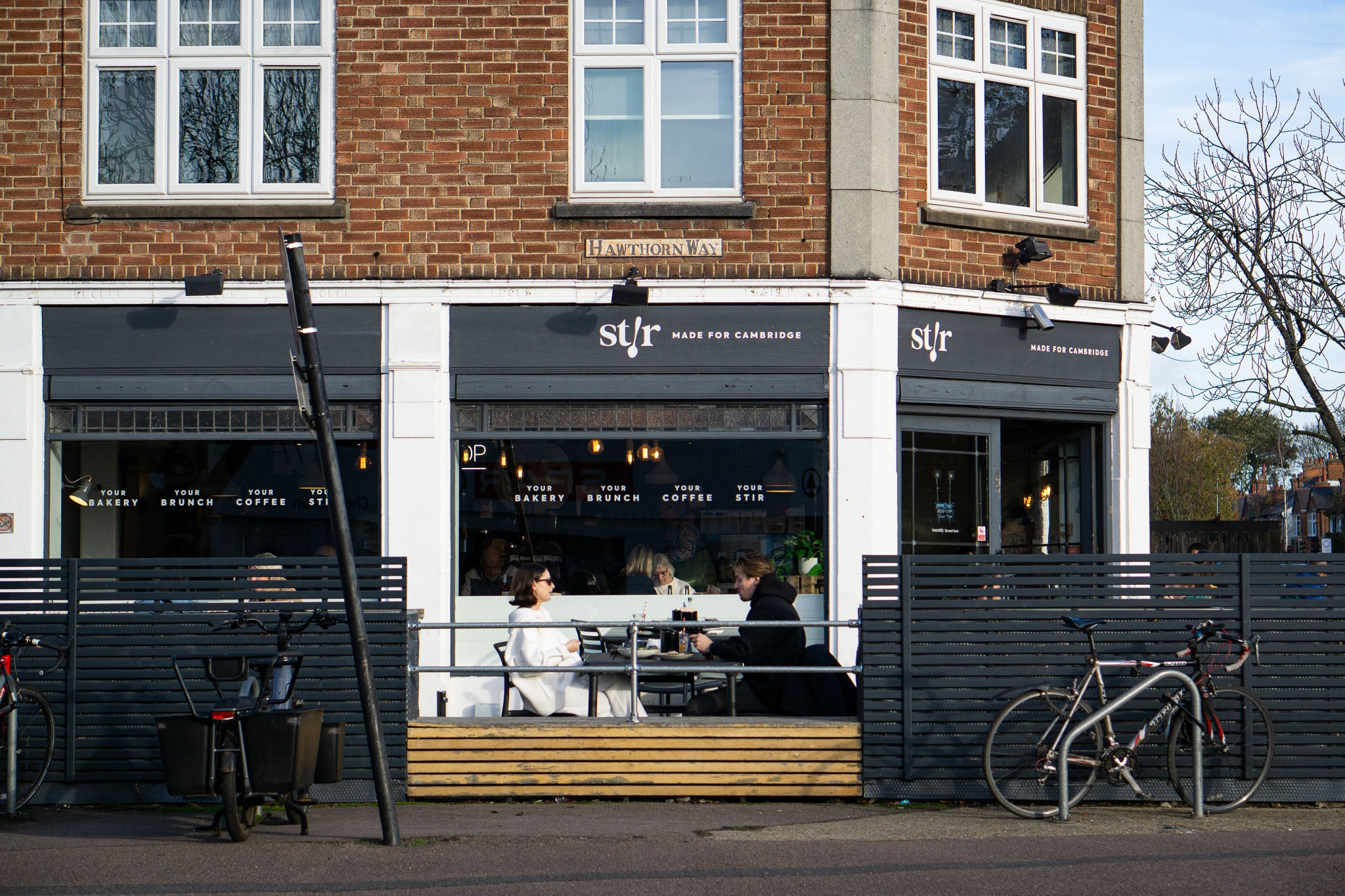 The Hawthorn Way corner storefront of Stir on Chesterton Road, with st/r MADE FOR CAMBRIDGE signage, pavement seating, bicycles parked outside and diners at an outdoor table