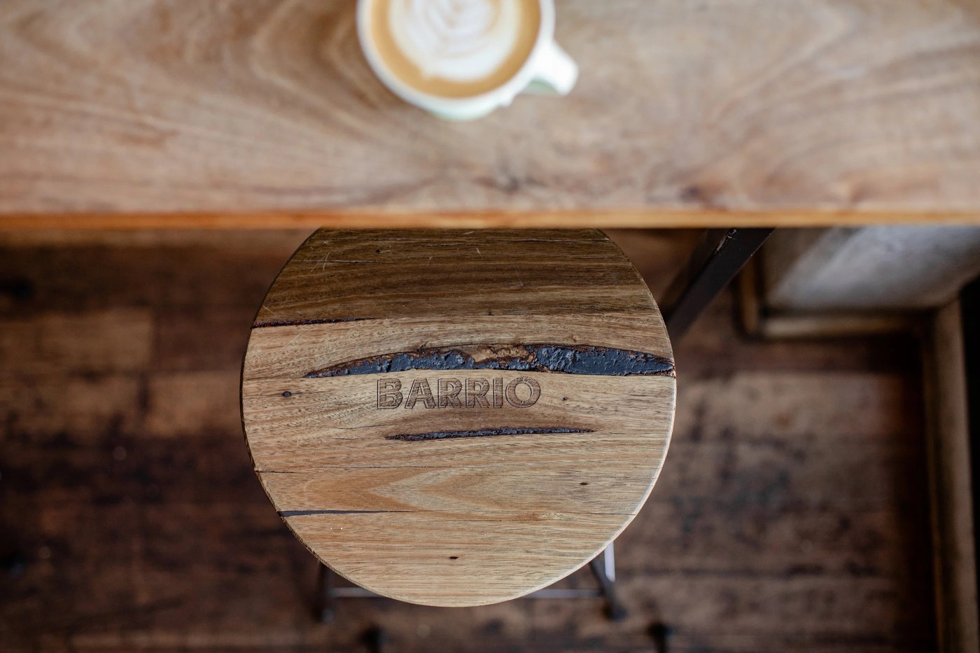 An overhead view of a wooden stool branded with the Barrio name beneath a table, with a latte visible on the tabletop above