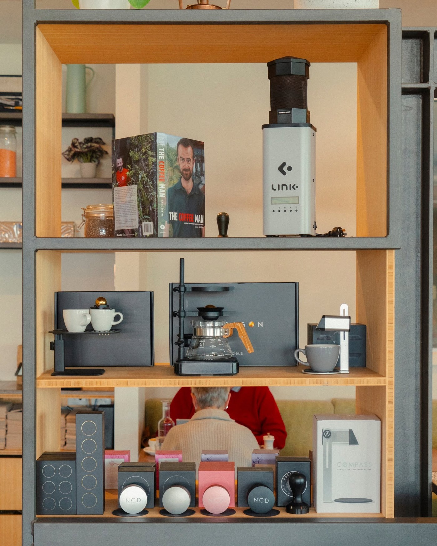 A shelving unit at ONA Coffee House stocked with a home coffee roaster, brewing gear, Sasa Sestic's book The Coffee Man, and retail accessories, with seating visible below