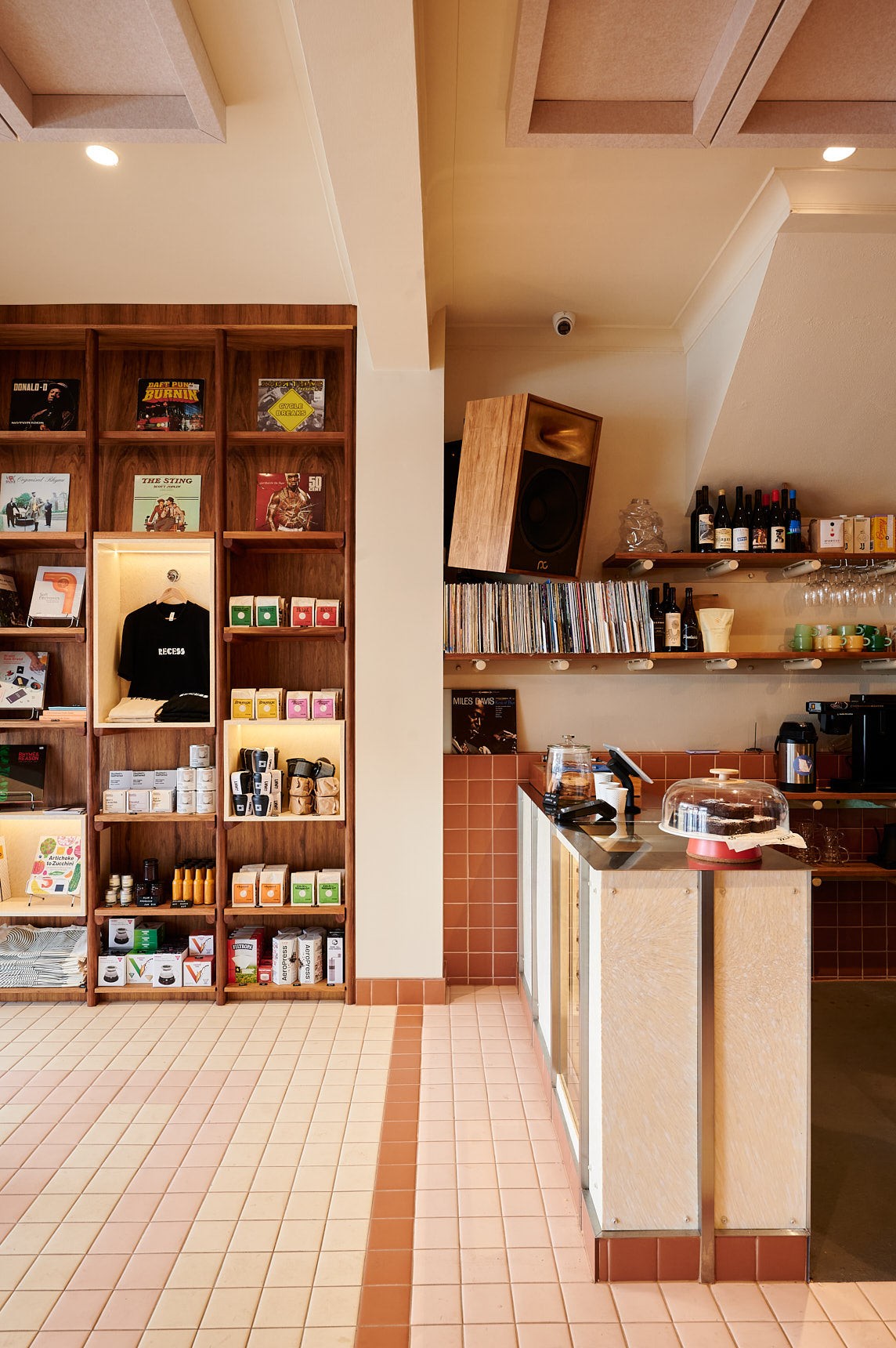 Retail shelving at Recess Coffee stocked with vinyl records, brew gear, and roasted bean bags, beside a terracotta tiled counter