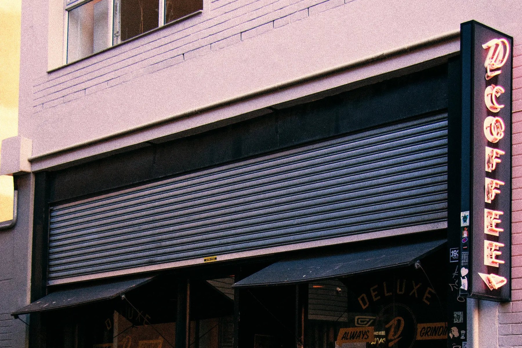 The Deluxe Coffeeworks shopfront on Kloof Street in Gardens at dusk, a vertical pink neon D COFFEE sign on the right and the DELUXE COFFEEWORKS window lettering above the entrance