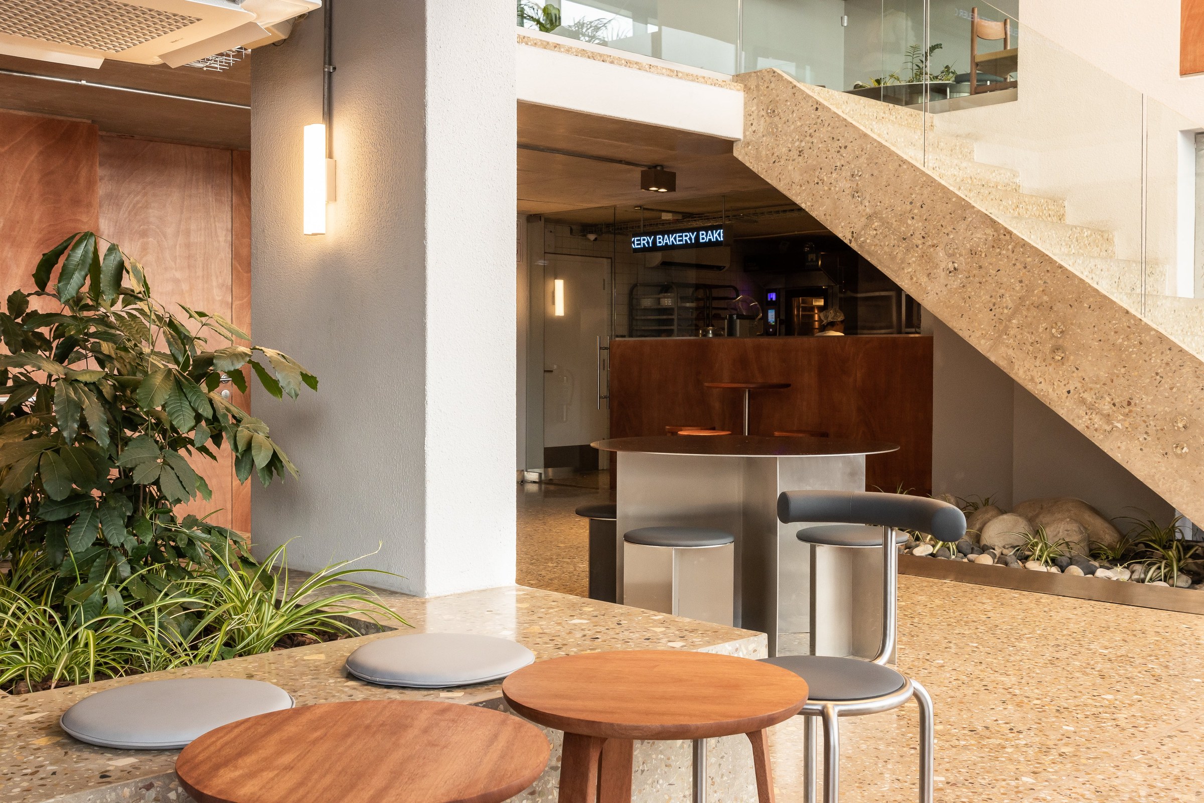 The ground floor of Espressolab De Waterkant with round timber and steel stools in front of a concrete and terrazzo staircase, planted beds and the glowing bakery sign behind