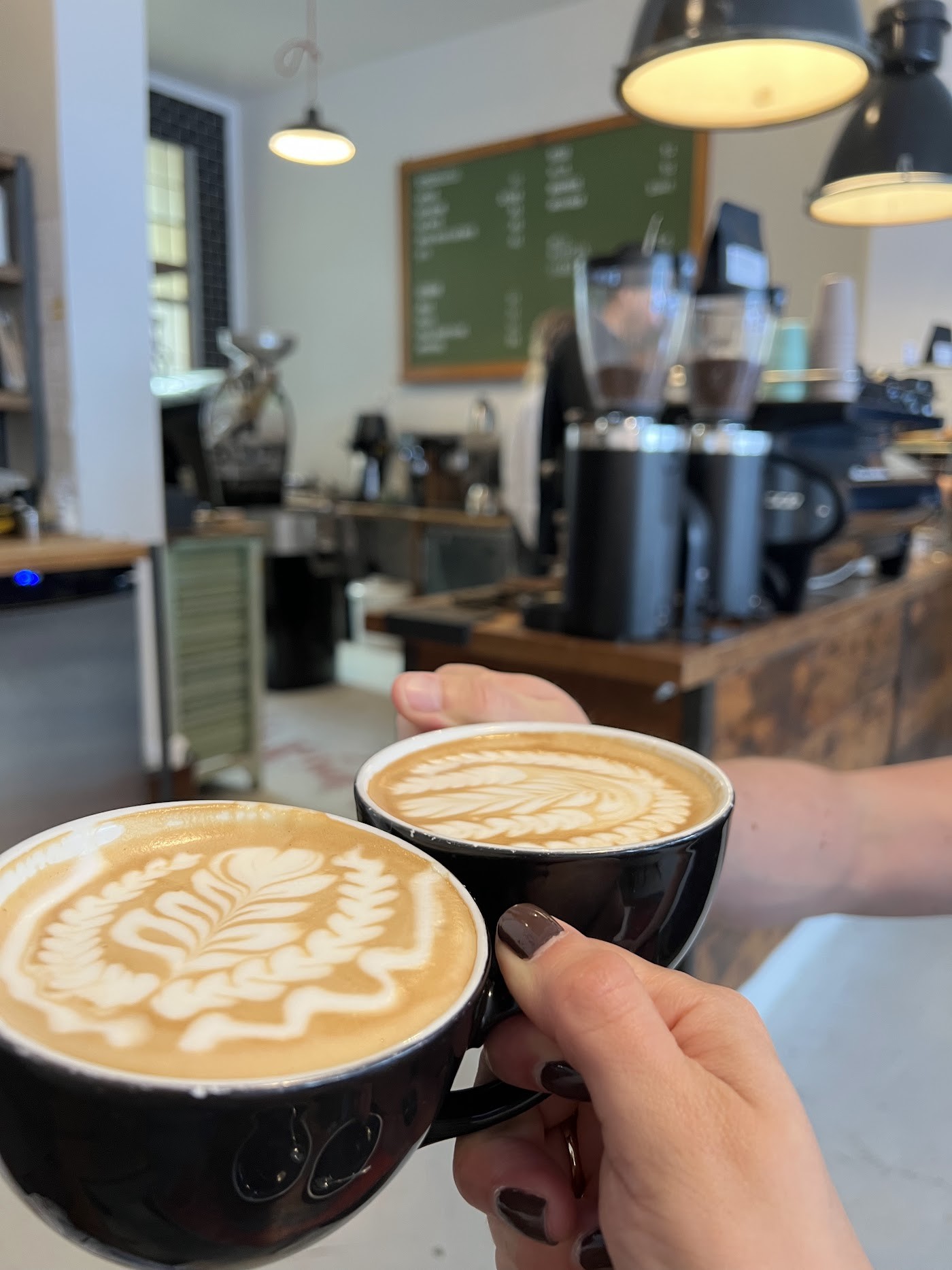Two black mugs of latte art coffee held up close to camera with a roastery counter, grinders and green chalkboard menu blurred in the background