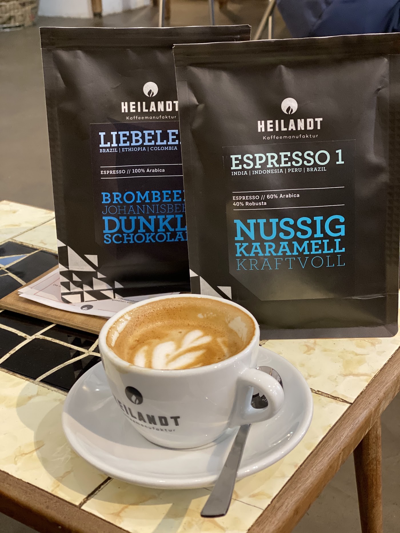 Two black bags of Heilandt coffee beans labeled Espresso 1 and Liebelei standing behind a white cup of latte art on a saucer with a spoon