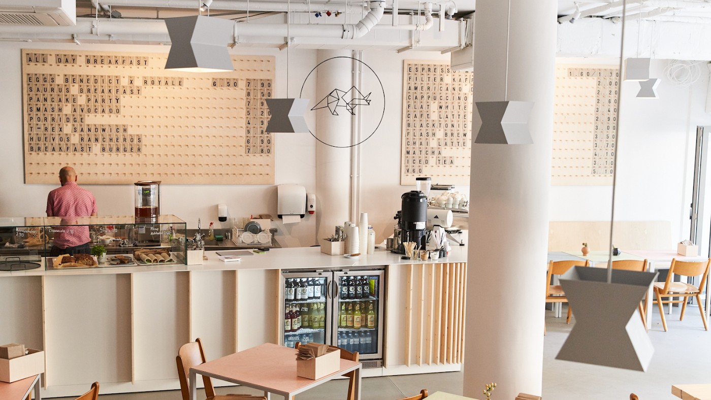 Interior of Kaffeesaurus with a wooden letterboard breakfast menu, a dinosaur skeleton logo on the wall, a coffee counter and pink cafe tables, one person facing away at the counter