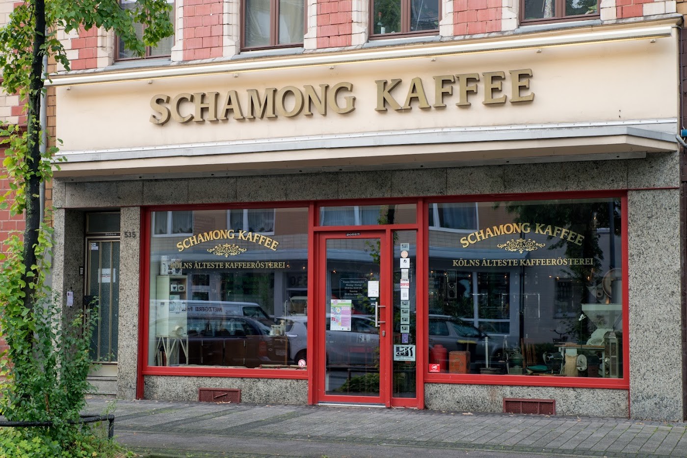 Storefront of Schamong Kaffee with gold lettering reading Schamong Kaffee Kölns älteste Kaffeerösterei above red framed windows and door, seen through green leaves