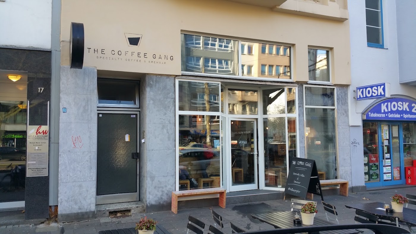 Cream colored storefront of The Coffee Gang with large glass windows, an outdoor bench and a sandwich board on the pavement, no people in view