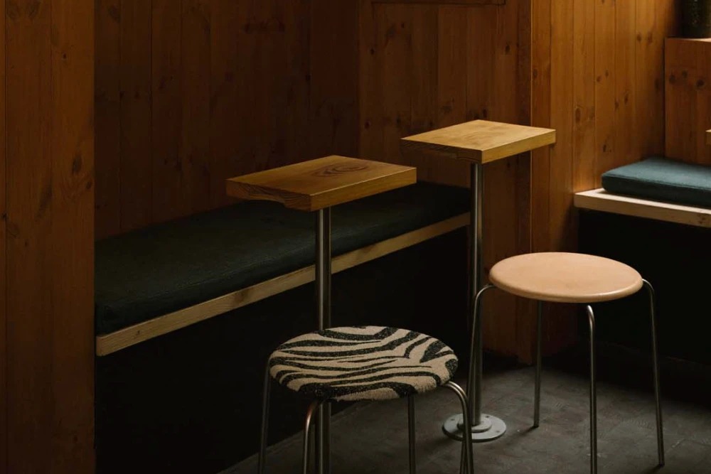 A pine lined seating nook at Cabin with a built in bench, small wall mounted tables, and two stools upholstered in leather and zebra patterned boucle