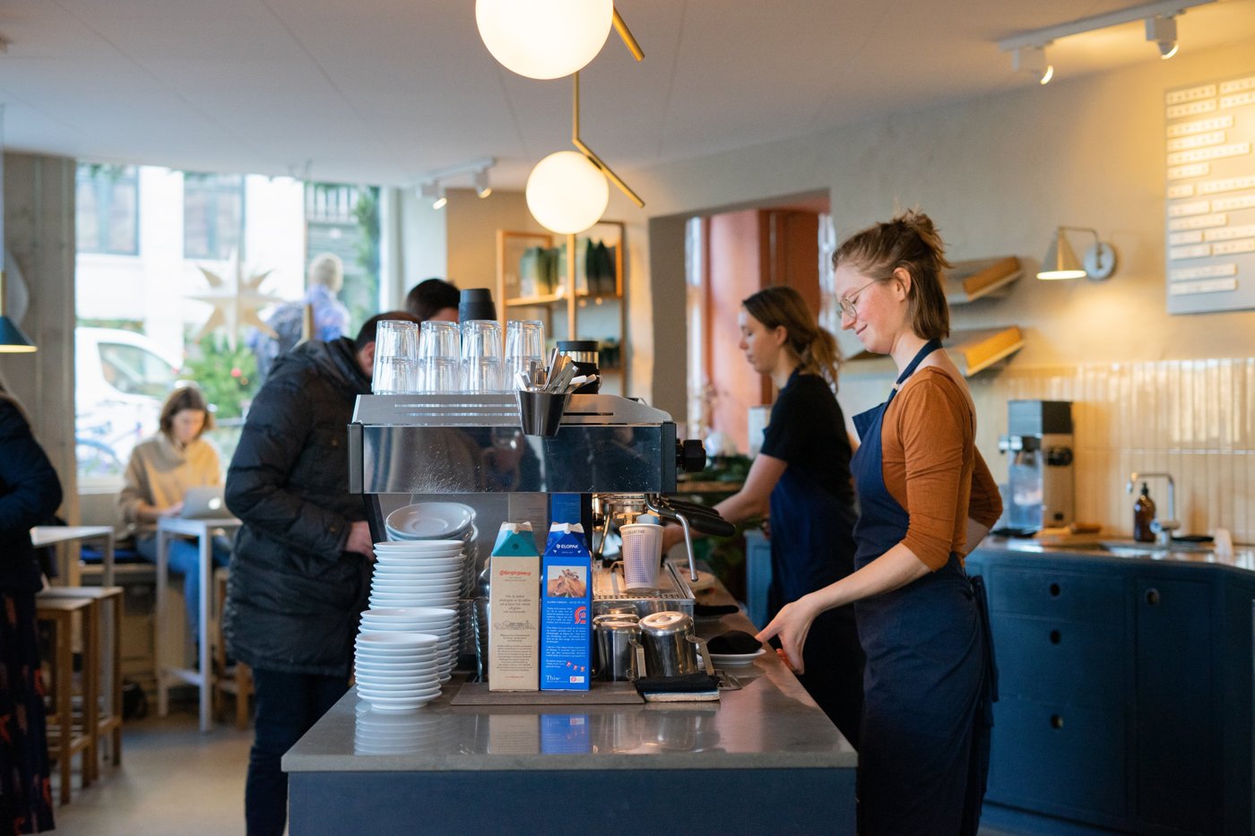 Baristas working behind the counter at Coffee Collective, globe pendant lights, glassware and blue-tiled shelving