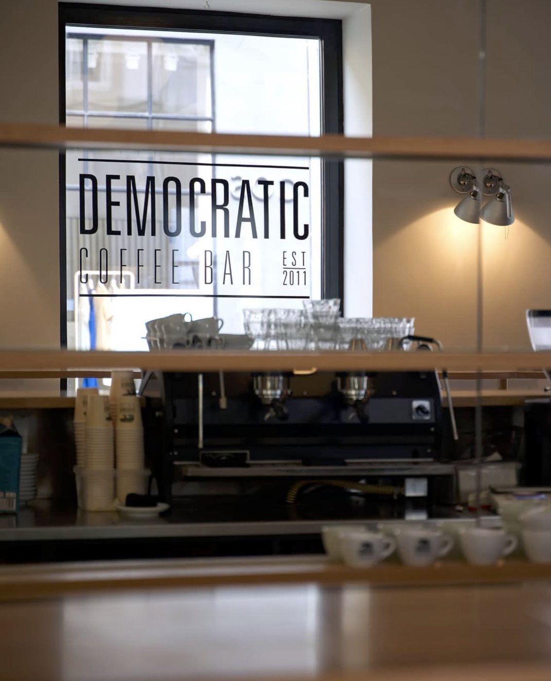 The Democratic Coffee Bar logo etched on the window behind the espresso machine and stacked cups