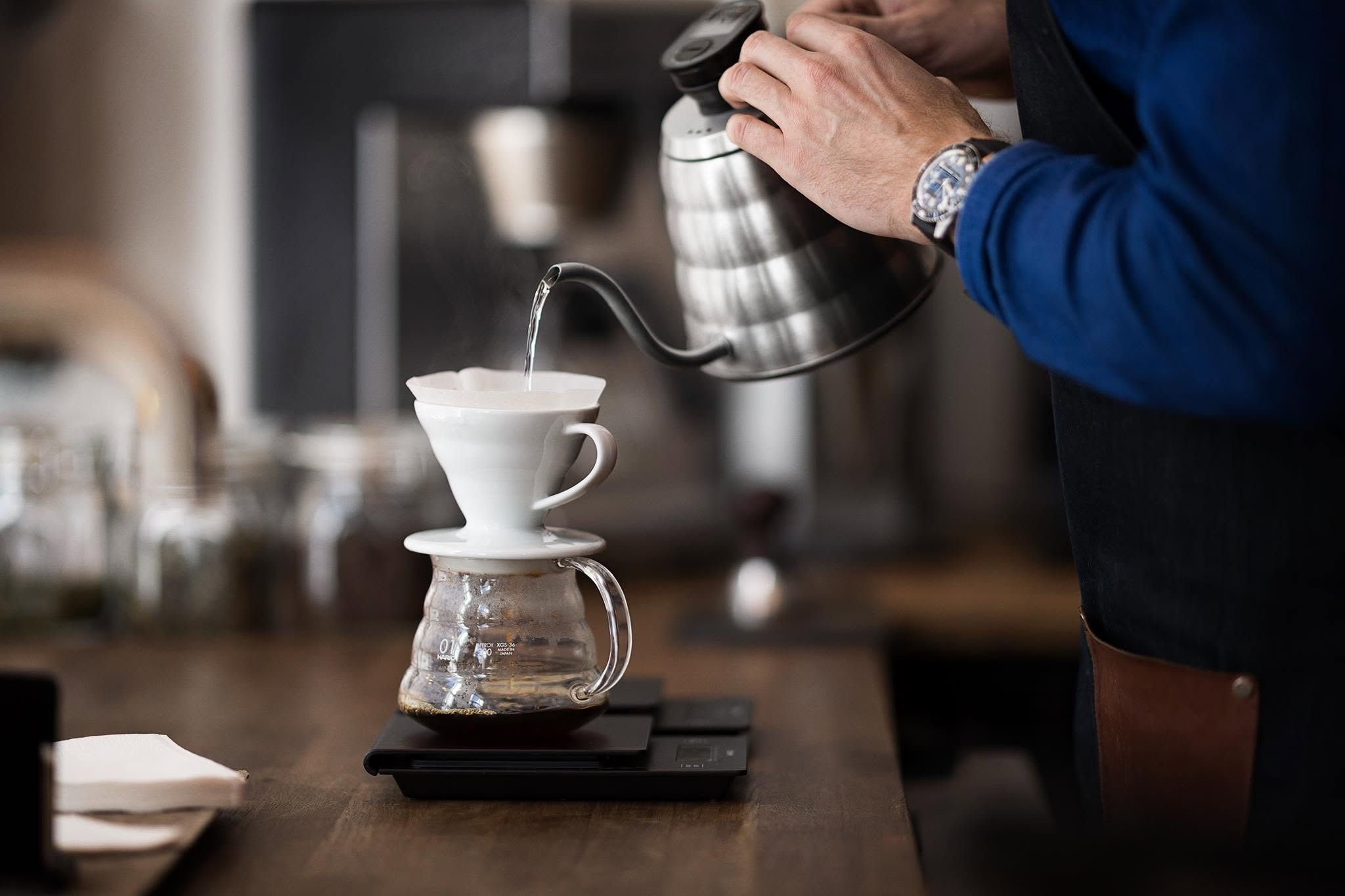 A barista's hands pouring hot water from a gooseneck kettle over a V60 dripper set on a digital scale, coffee brewing below