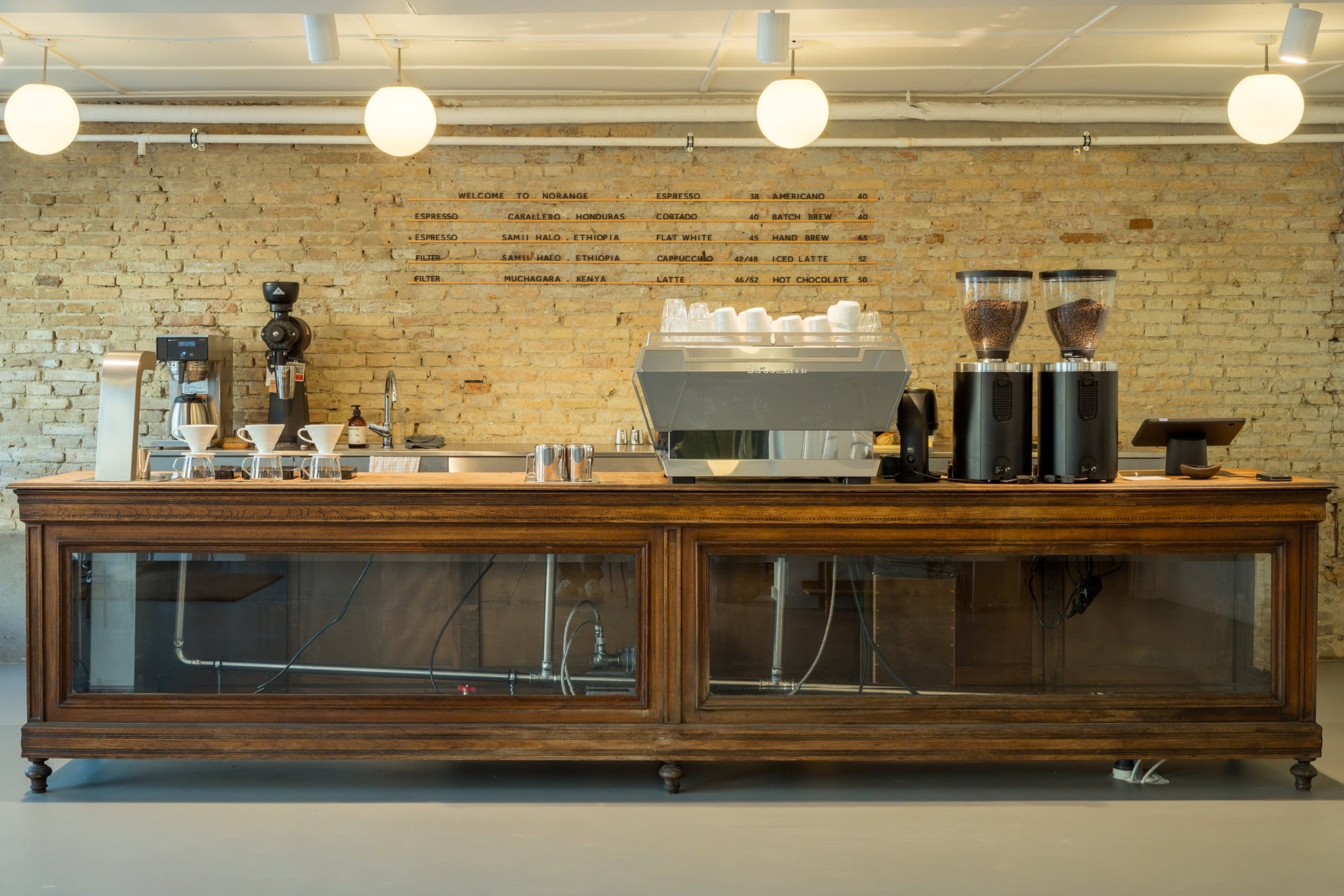 Norange's antique wooden counter beneath a brass menu board on exposed brick, with an espresso machine and two bean grinders