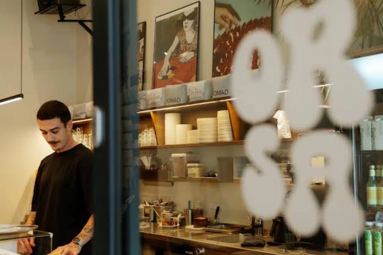 A barista working the counter at Orsa, seen through the window past the ORSA decal, with vintage style art prints and a retail drinks fridge behind