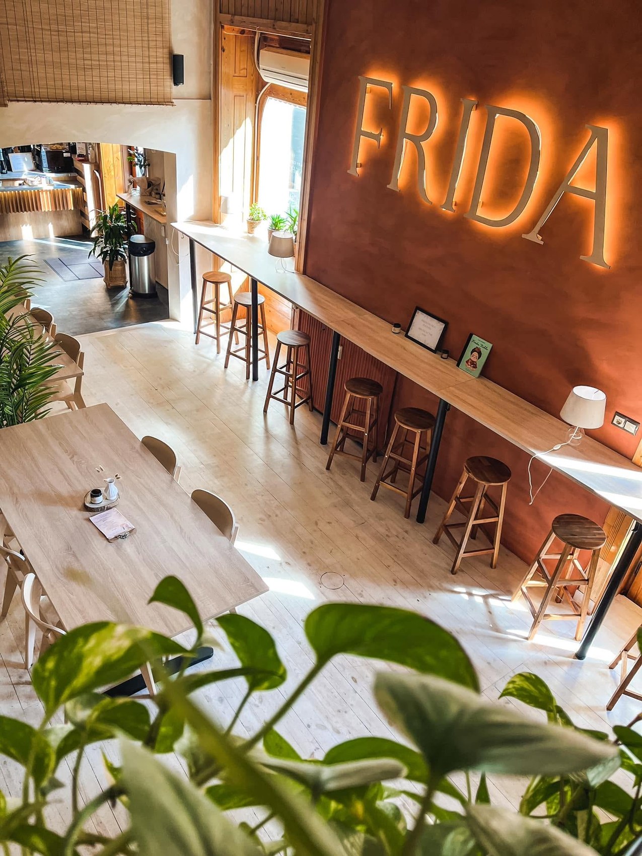 The backlit FRIDA sign on a terracotta wall above a long bar counter with stools, and a wooden communal table with potted plants in the foreground