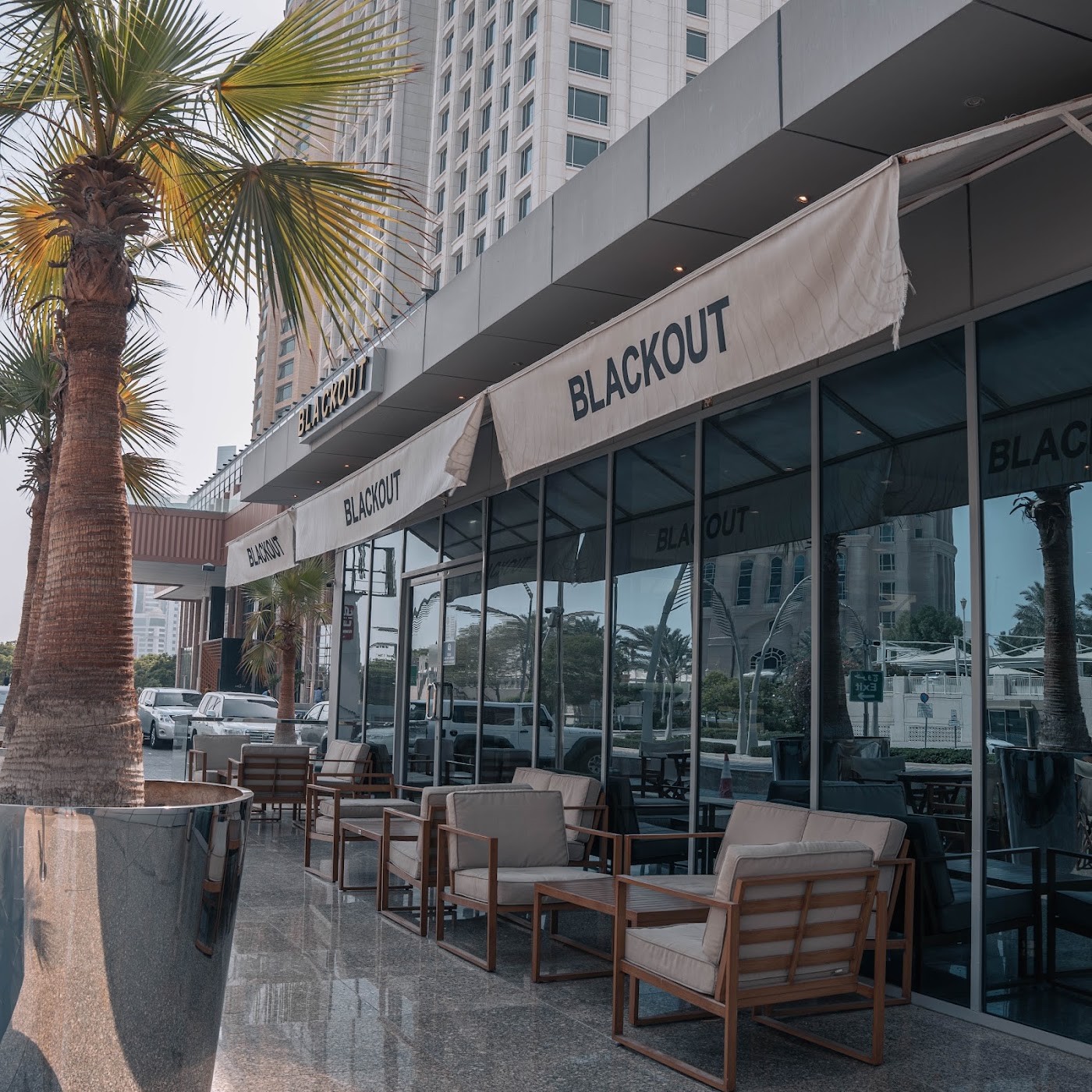 The Blackout storefront awning and glass facade beneath a palm lined terrace with wood framed lounge seating