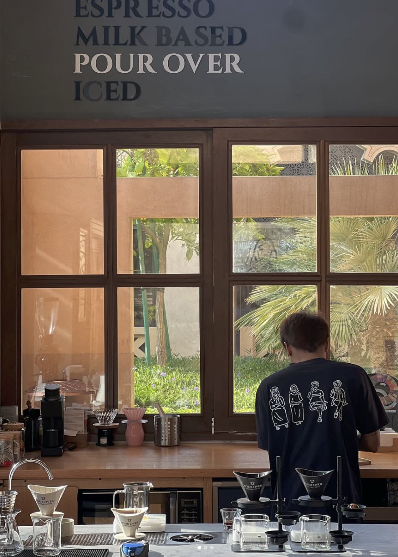 A barista working at the pour over station beneath a wall menu reading Espresso, Milk Based, Pour Over, Iced, with palm trees visible through the window