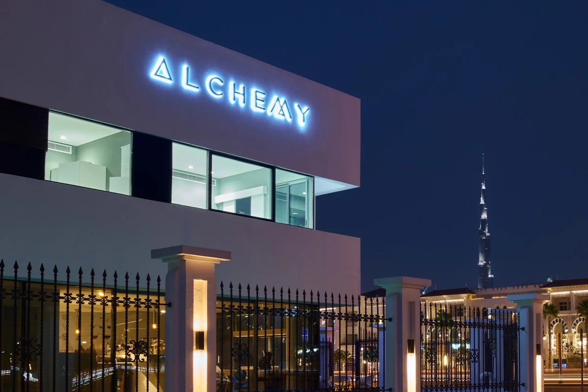 The glowing blue ALCHEMY sign on the white villa at night with the Burj Khalifa rising behind the iron fence