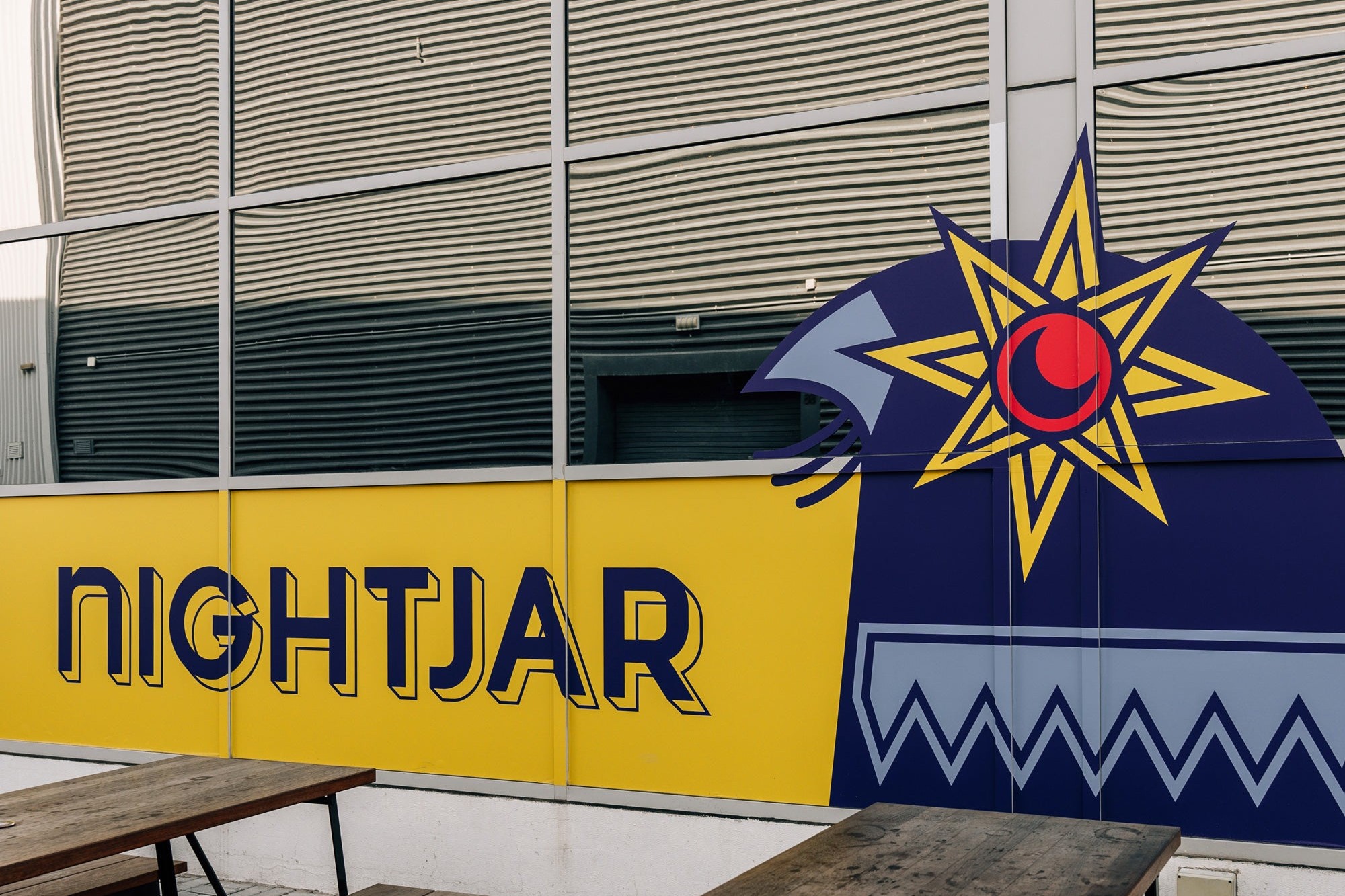 The yellow and navy Nightjar mural with its star eyed night bird above wooden picnic tables outside the Alserkal Avenue warehouse