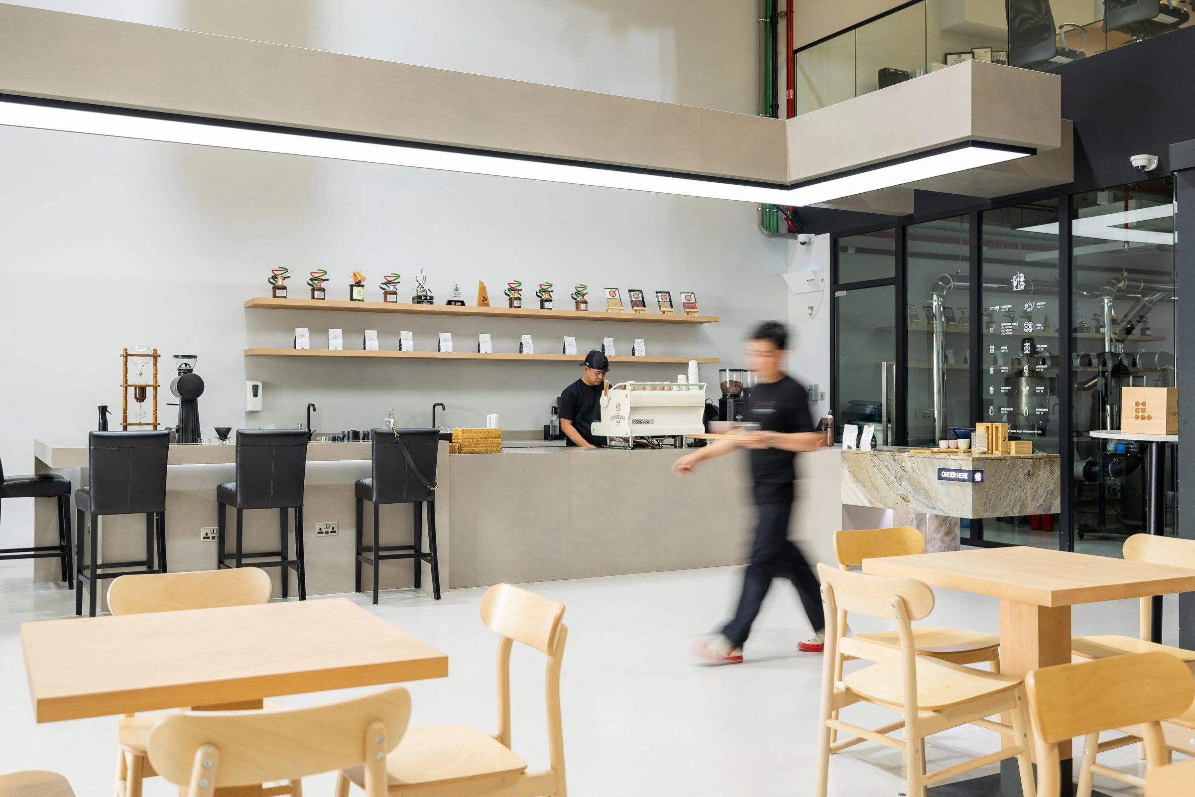 Inside The Espresso Lab roastery, a coffee bar with a shelf of trophies and bagged beans, wooden tables and the glass walled roasting room behind