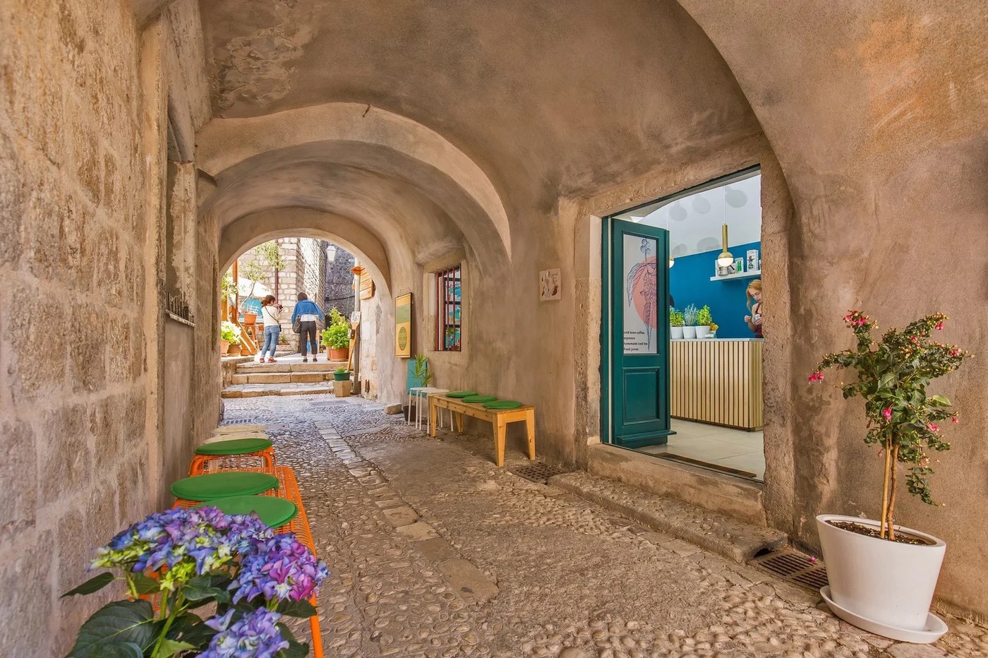 The stone tunnel entrance to Cogito Coffee Shop in Dubrovnik's Old Town, with a teal door open to the counter and green benches lining the passage