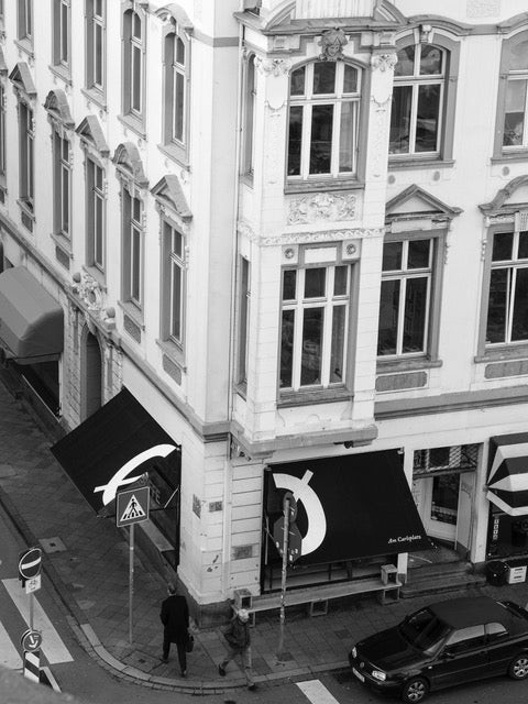 An aerial black and white view of the historic corner building housing Cøffe am Carlsplatz, its Cø logo printed on two black awnings above the street