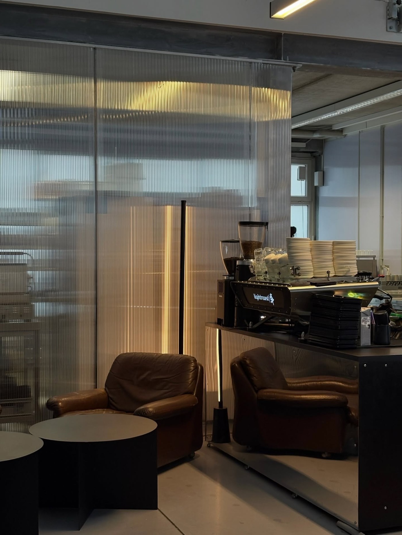 The Lightroast branded espresso machine and grinder beside stacked cups, with leather armchairs and a corrugated glass partition in the seating area