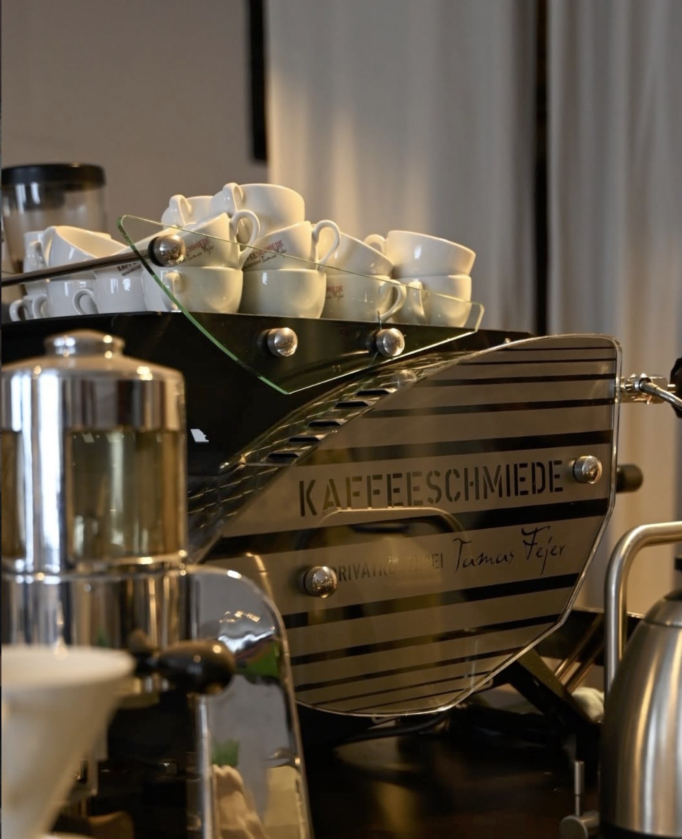 The KAFFEESCHMIEDE Privatrösterei Tamas Fejer espresso machine with a stack of branded cups on top