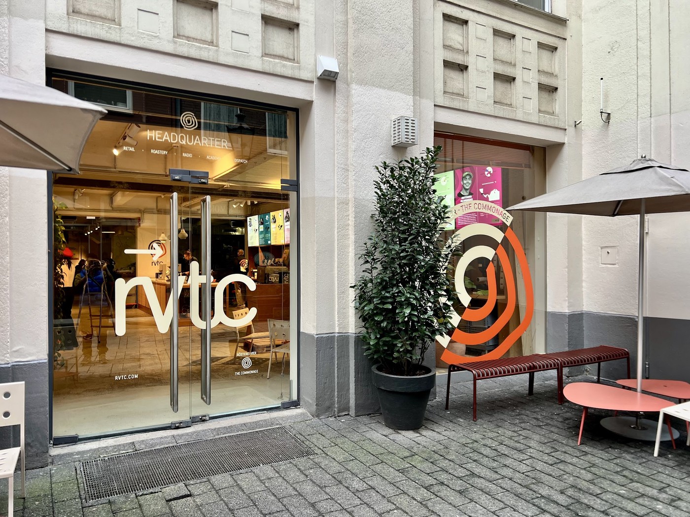 The glass storefront of RVTC Rösterei Vier with its logo on the door, an orange spiral mural, potted plants, and outdoor benches and tables on the Altstadt pedestrian street