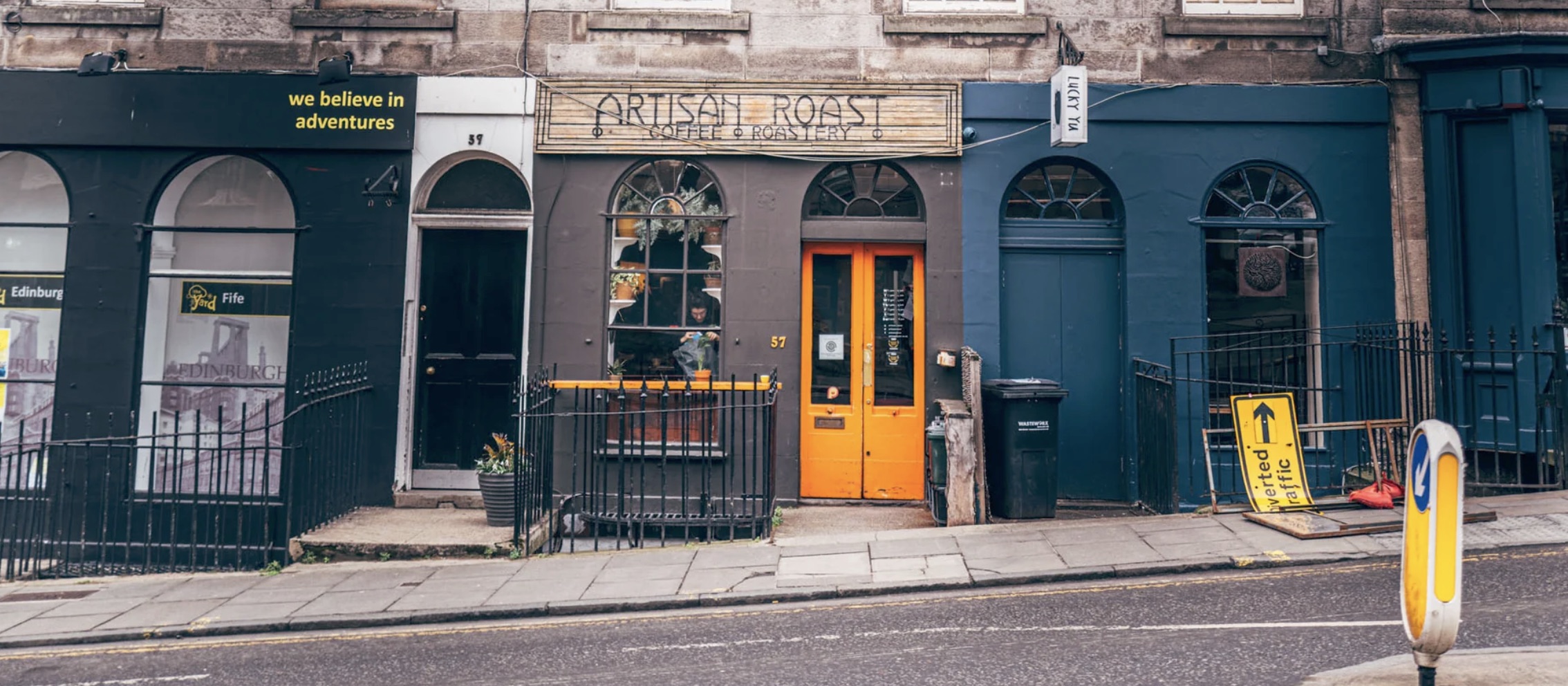 Artisan Roast's dark grey Broughton Street shopfront with a carved ARTISAN ROAST COFFEE ROASTERY sign and a bright orange double door at number 57, flanked by arched windows on a stone tenement