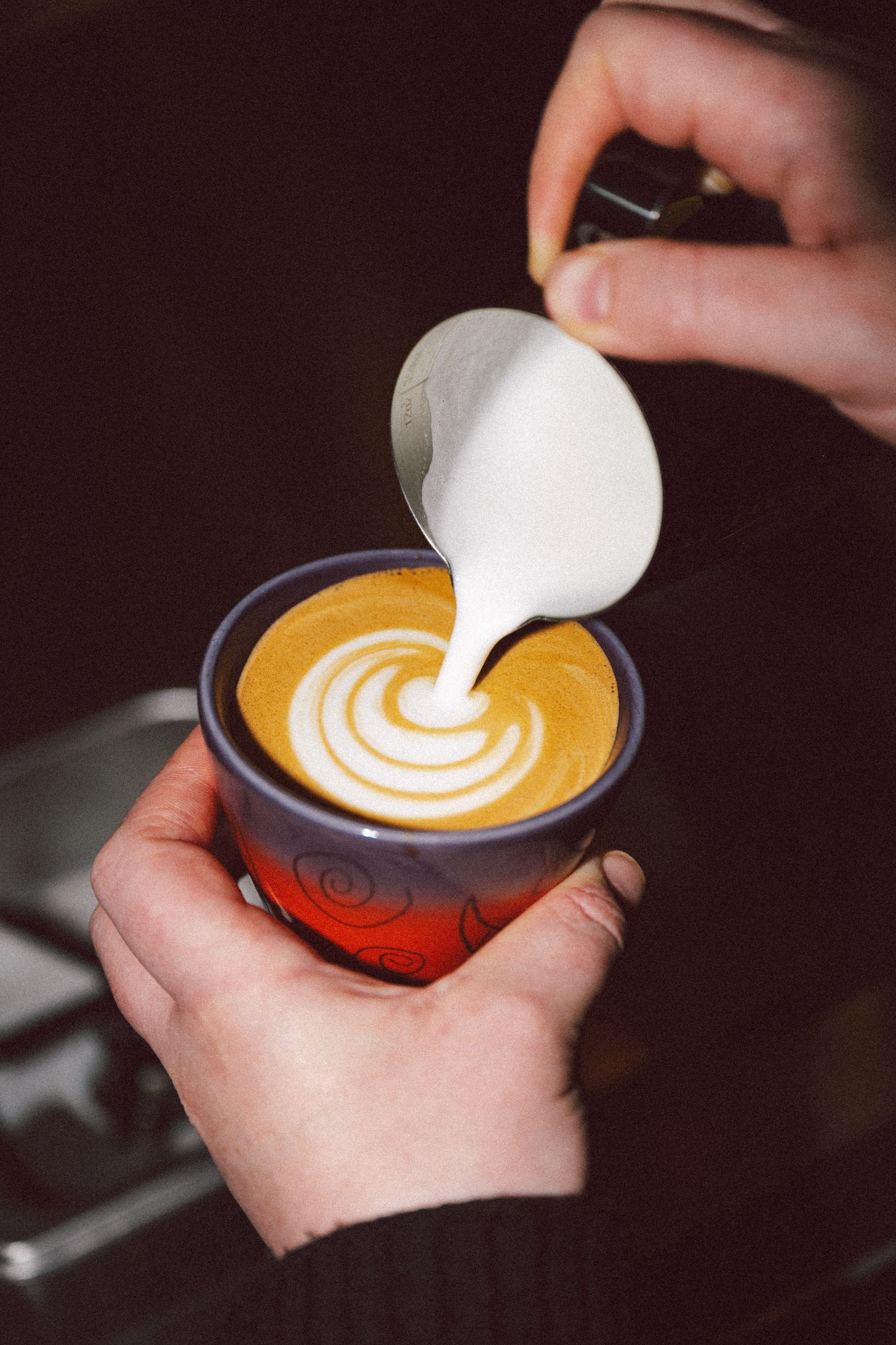 A barista pouring steamed milk from a jug into a purple and red cup, drawing a rosetta of latte art