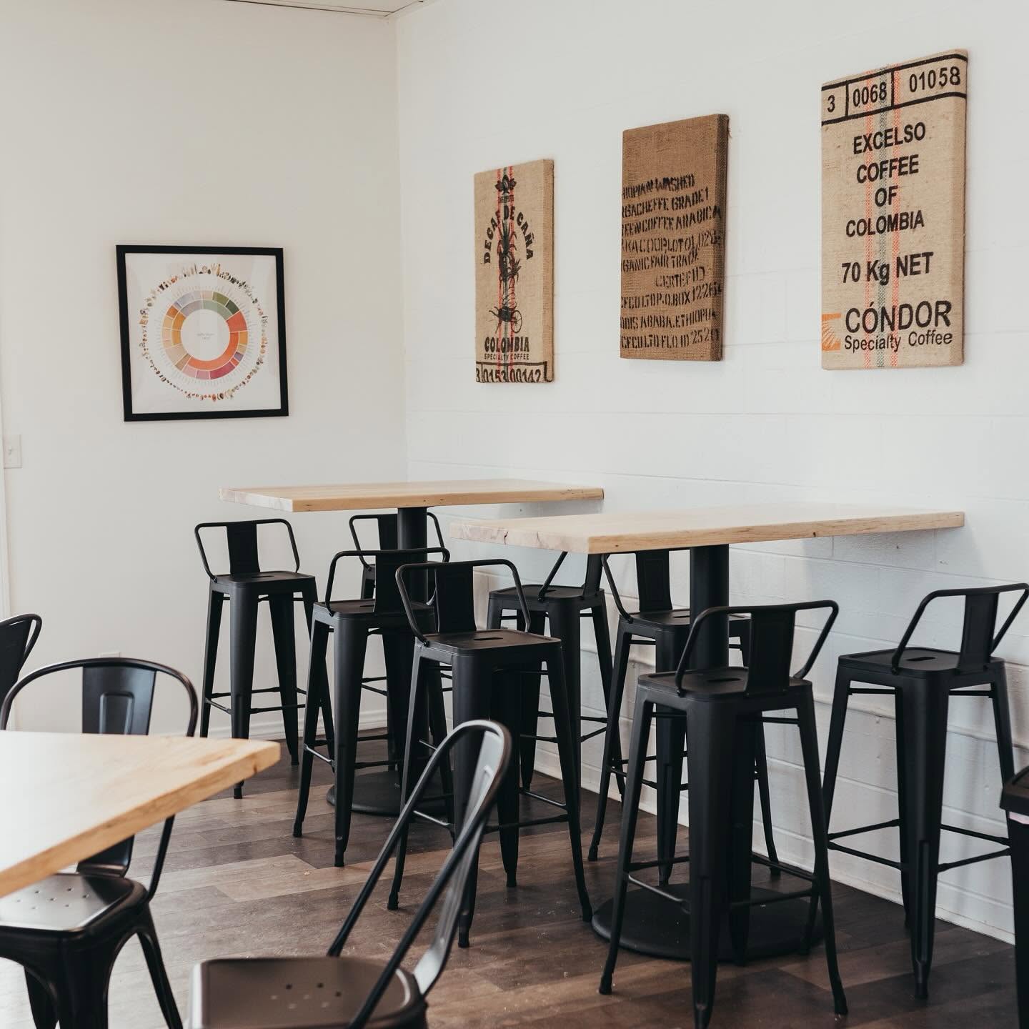 Bedrock's minimalist interior with light wood high tables and black metal stools against a white wall hung with a coffee flavor wheel and framed burlap coffee sacks