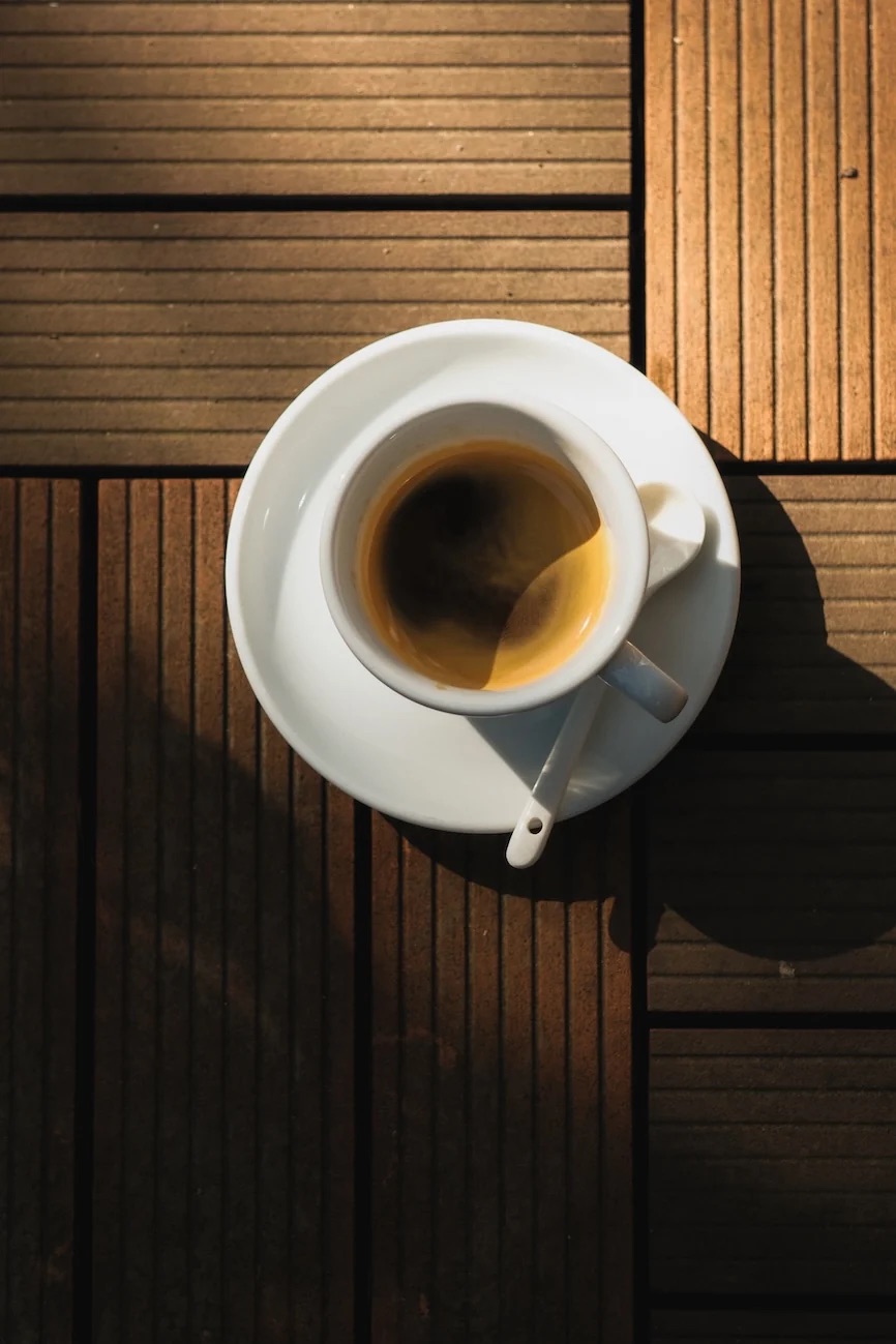 A top-down espresso in a white cup and saucer with a spoon resting on the rim, on a sunlit wooden deck