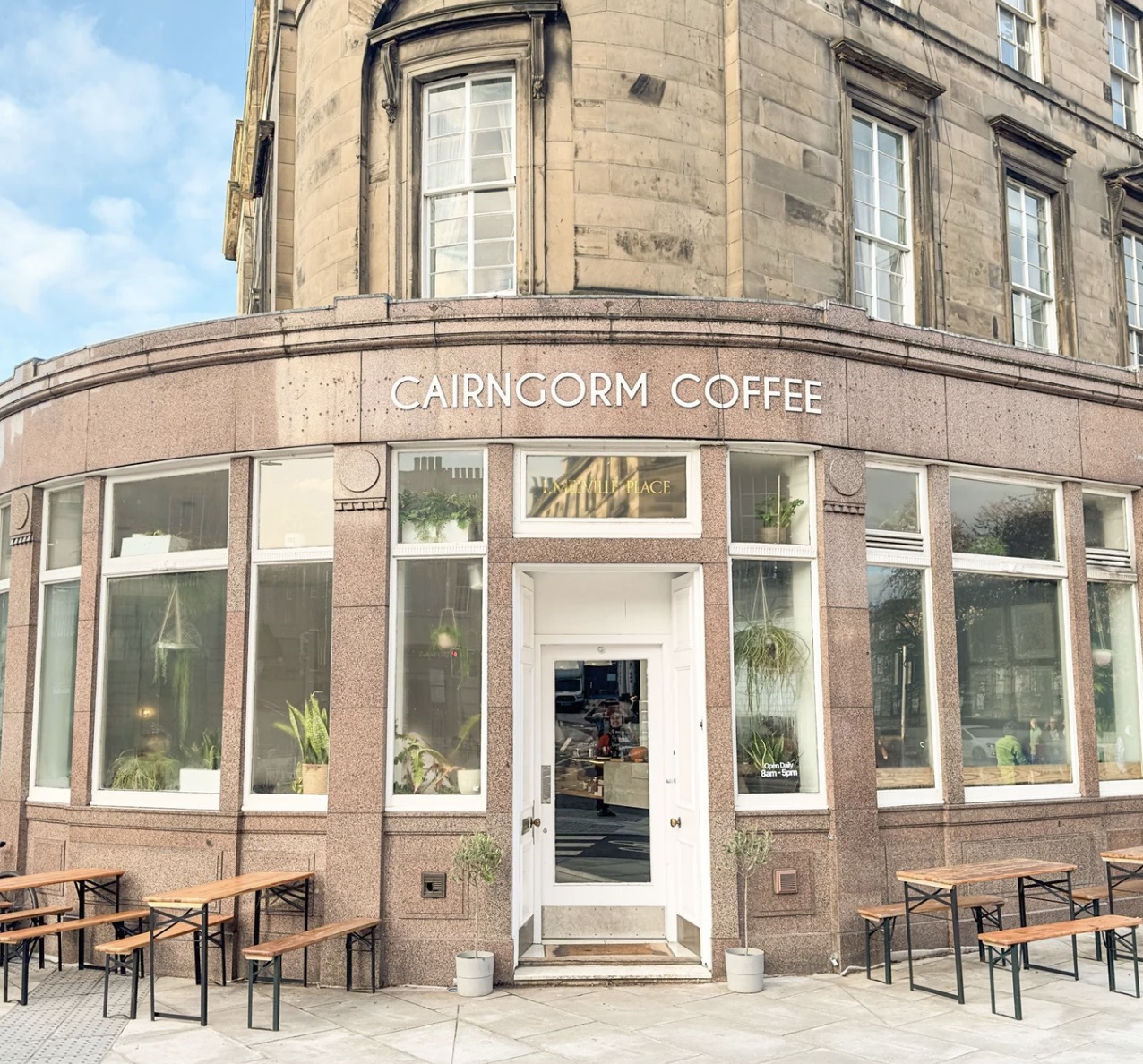 The rounded stone corner frontage of Cairngorm Coffee on Melville Place, with CAIRNGORM COFFEE lettering above tall plant-filled windows and wooden bench seating on the pavement
